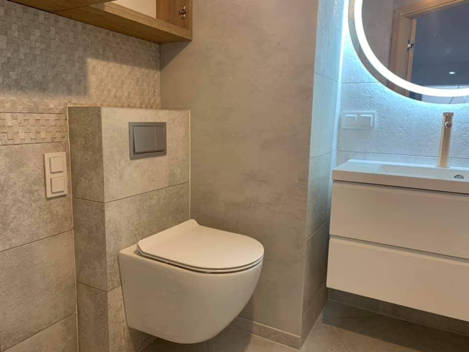 Nowoczesna łazienka z podwieszaną toaletą, okrągłym lustrem z podświetleniem LED i szafką z umywalką w odcieniach bieli i szarości, płytki imitujące kamień na ścianach.