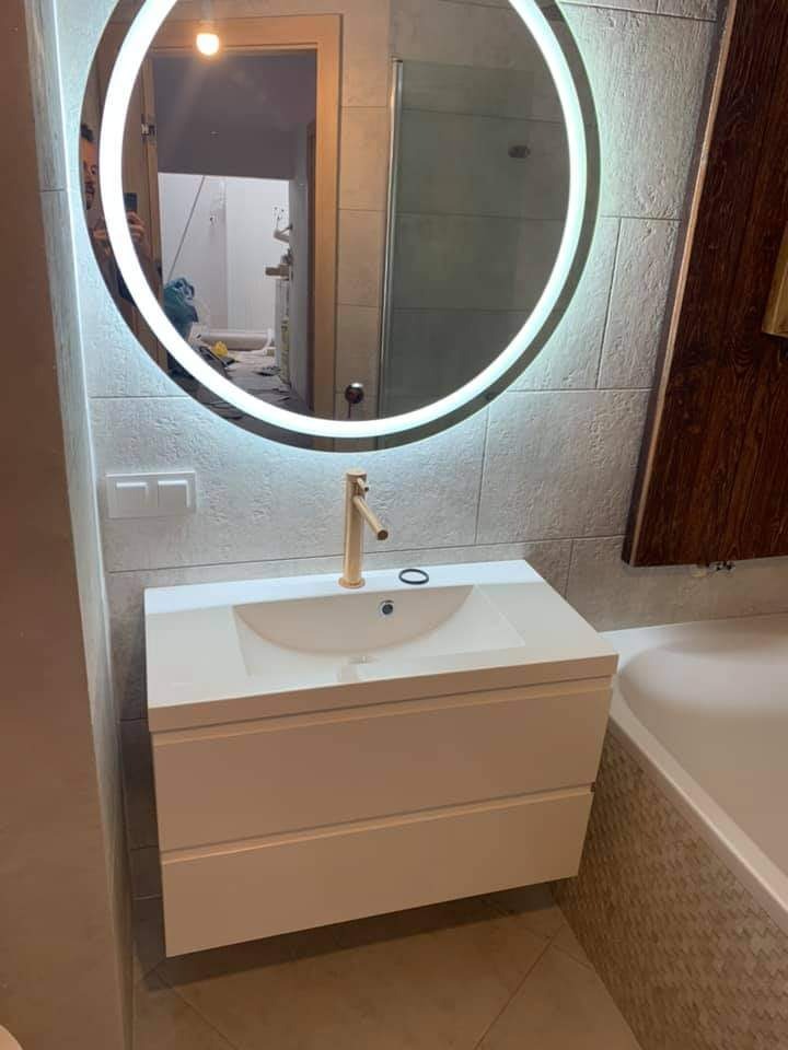 Biała szafka pod umywalkę z szufladami, złota bateria i okrągłe lustro z podświetleniem LED w nowoczesnej łazience z płytkami imitującymi kamień.