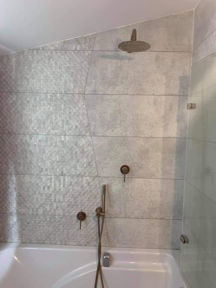 Nowoczesna łazienka z wanną, szarą mozaiką i płytkami imitującymi beton, armatura w kolorze miedzi, deszczownica zamontowana na skośnej ścianie.