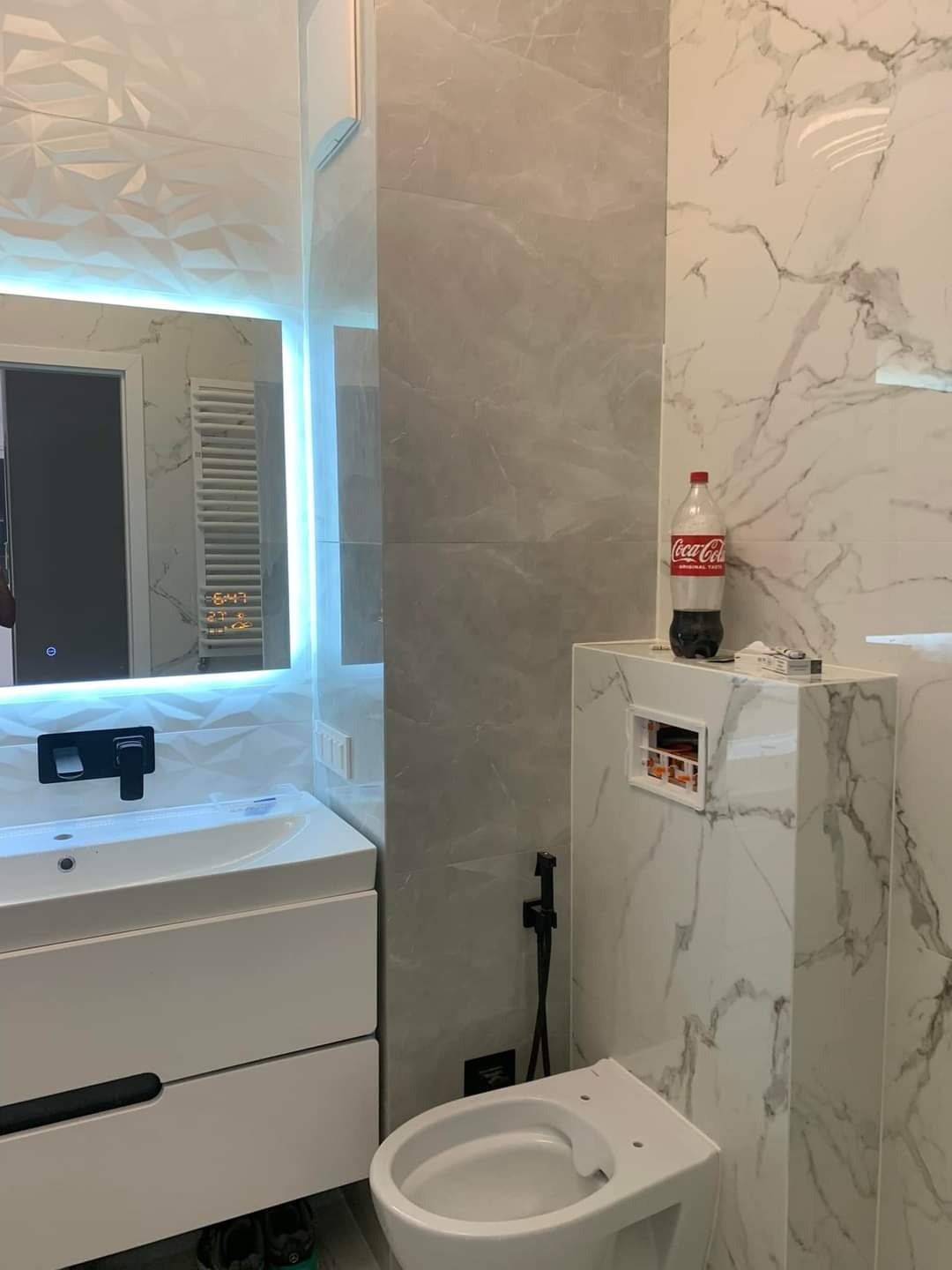Nowoczesna łazienka z marmurowymi płytkami, podświetlanym lustrem z wyświetlaczem, białą umywalką z szafką, toaletą i butelką Coca-Coli na obudowie stelaża podtynkowego.