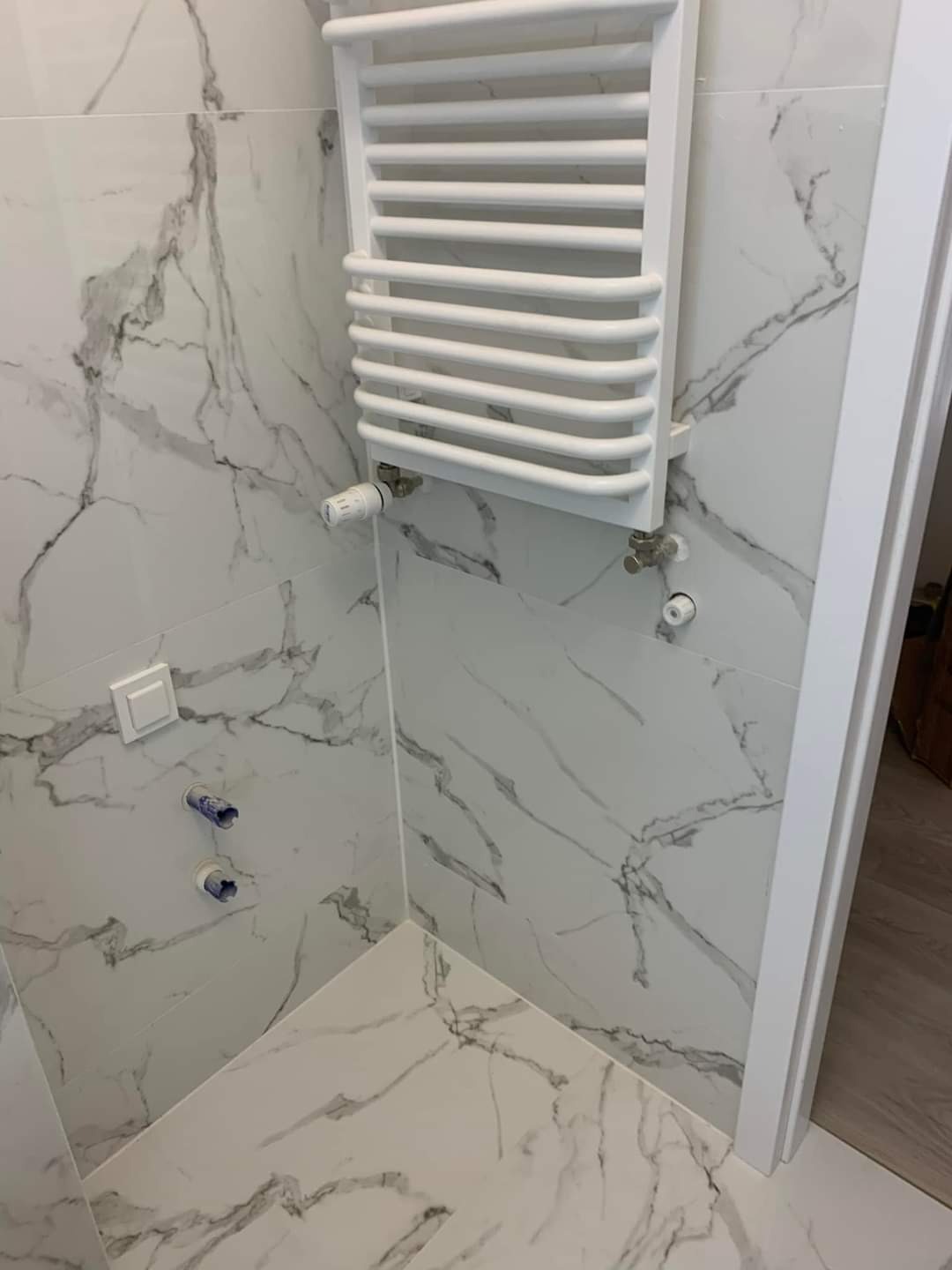 Nowoczesna łazienka wykończona białymi płytkami imitującymi marmur, z grzejnikiem drabinkowym i widocznymi przyłączami hydraulicznymi w trakcie montażu.