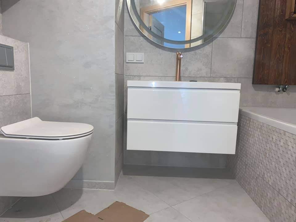 Nowoczesna łazienka z wiszącą miską WC, szafką pod umywalką z baterią w kolorze miedzi i okrągłym lustrem z podświetleniem LED, wykończona szarymi płytkami o zróżnicowanej fakturze.