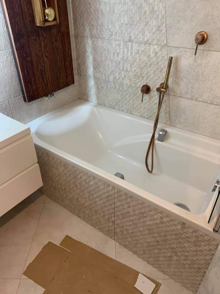 Biała wanna obudowana mozaiką w odcieniach beżu i szarości, złota bateria, płytki imitujące cegiełki na ścianie, drewniana półka z dekoracją, fragment kartonu na podłodze.