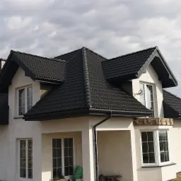 Nowoczesny dom jednorodzinny z ciemną dachówką ceramiczną, widoczne rynny i obróbki blacharskie, na podjeździe zaparkowany bus firmy dekarskiej.