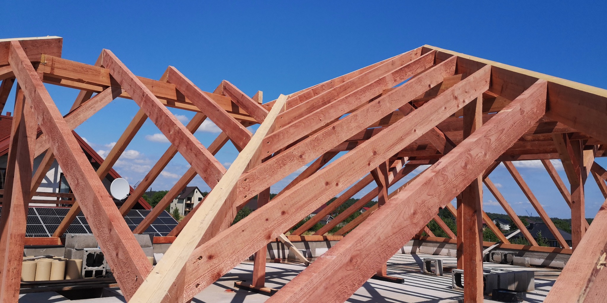 Konstrukcja drewniana dachu w trakcie budowy, widoczne elementy więźby dachowej z jasnego drewna na tle błękitnego nieba i zabudowań z panelami słonecznymi.