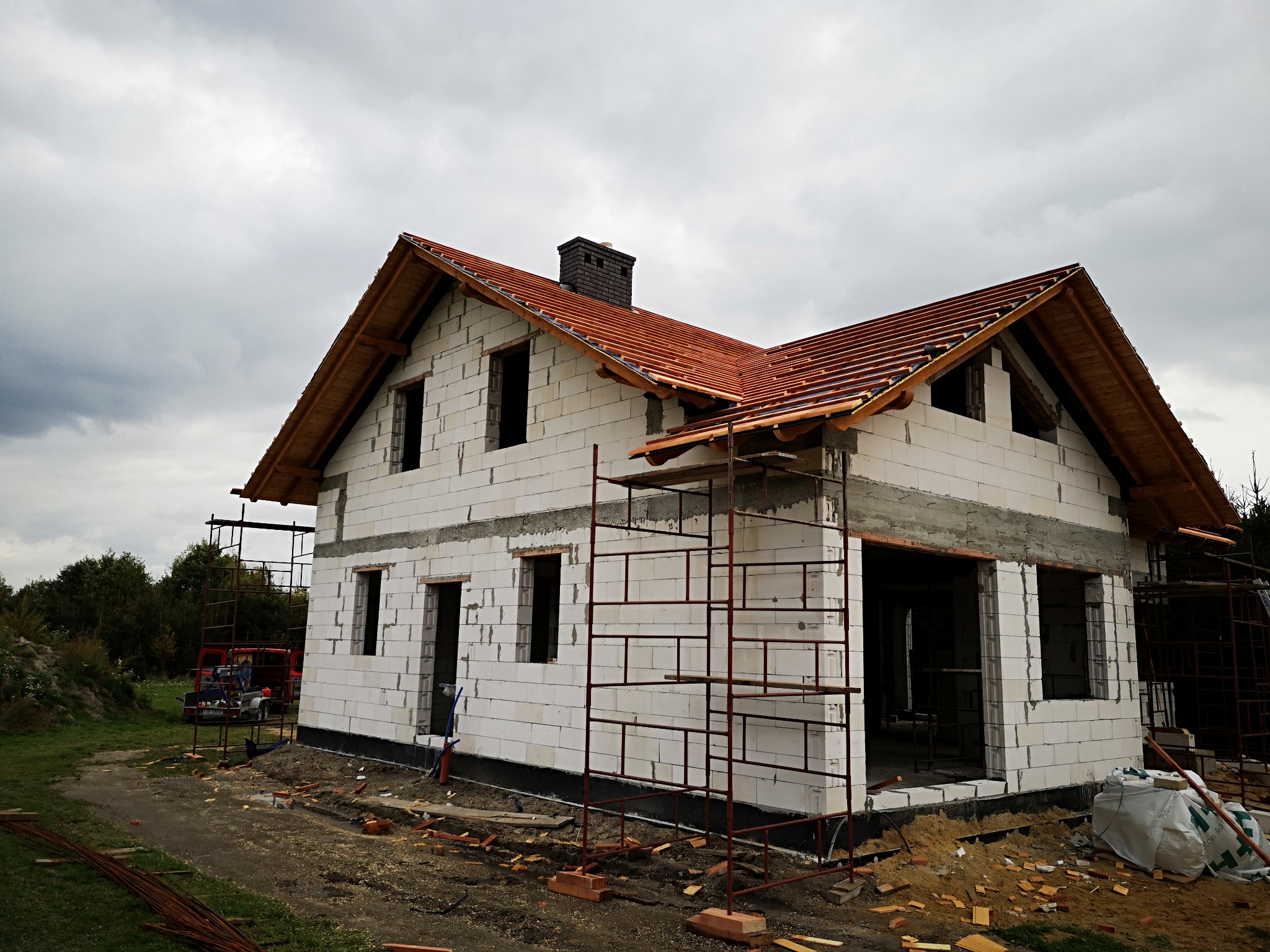 Surowy stan domu jednorodzinnego z białego bloczka, z częściowo ułożoną konstrukcją dachu z czerwonej dachówki oraz widoczną więźbą dachową pod pochmurnym niebem.