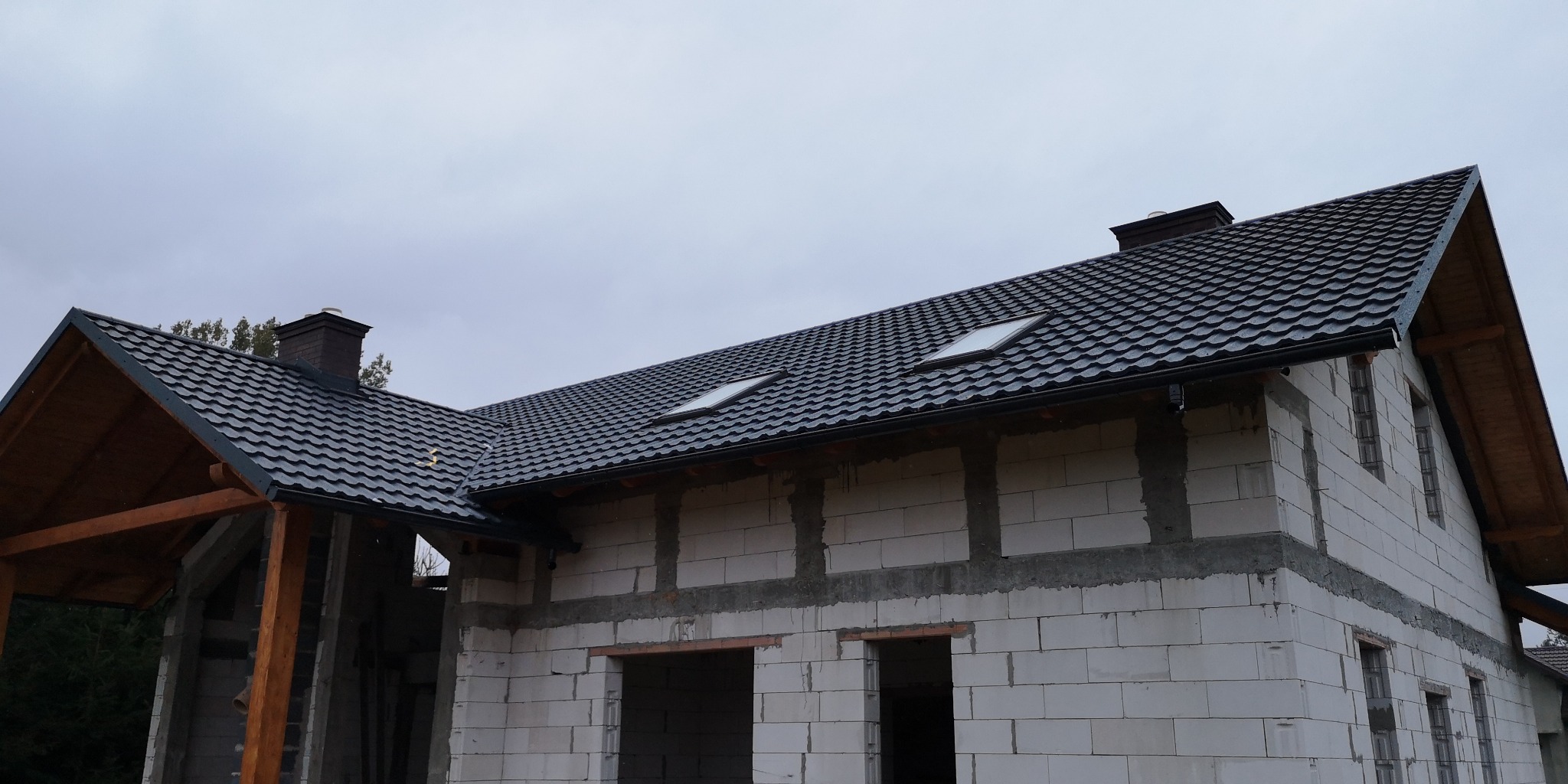 Dom w trakcie budowy z nowym, grafitowym dachem z dwoma oknami dachowymi i zamontowanymi rynnami, widoczne bloczki z betonu komórkowego.