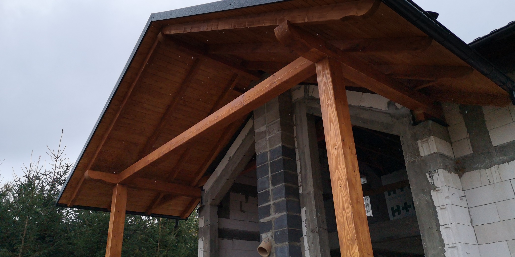 Drewniana konstrukcja z zadaszeniem w trakcie budowy, widoczne elementy więźby dachowej, rynna i surowy stan budynku z bloczków.