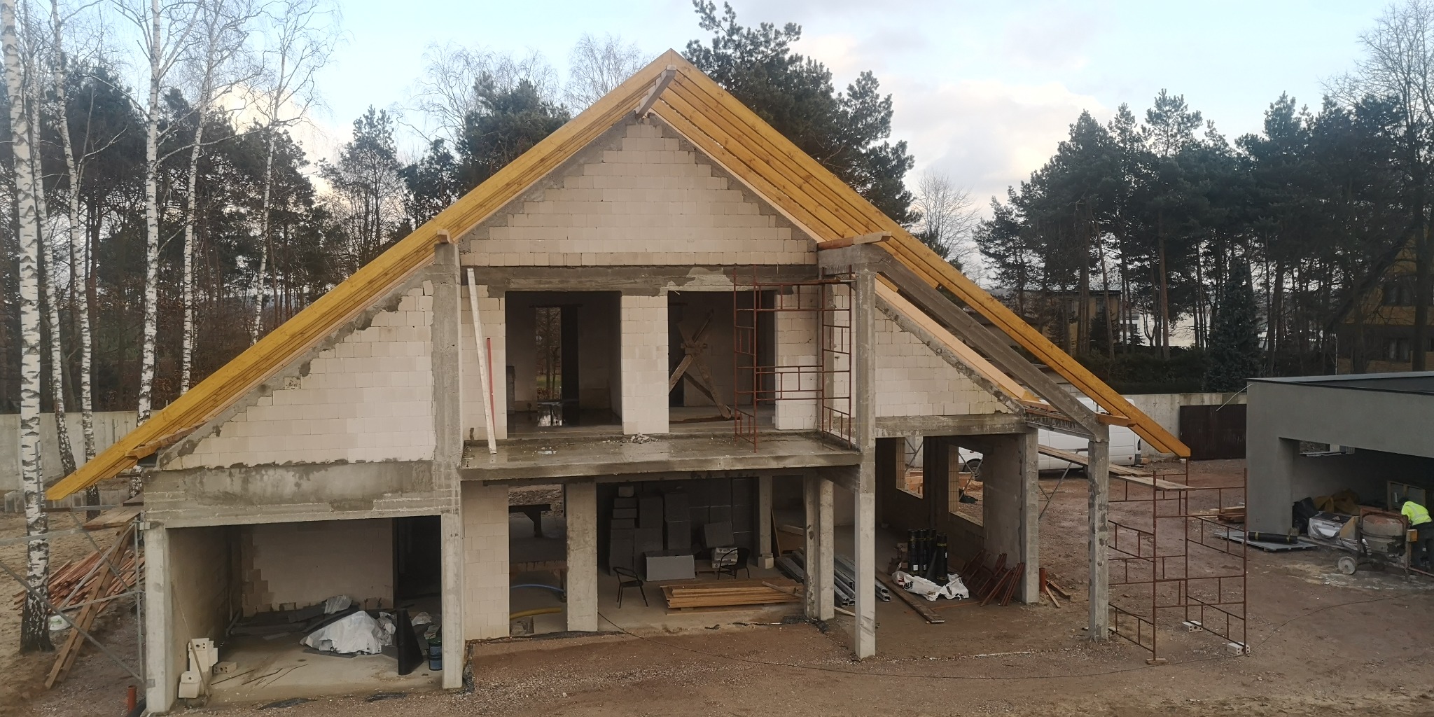 Surowy stan domu jednorodzinnego w trakcie budowy, z widoczną konstrukcją więźby dachowej z drewna, bloczki betonowe ścian, rusztowania oraz otaczający las.