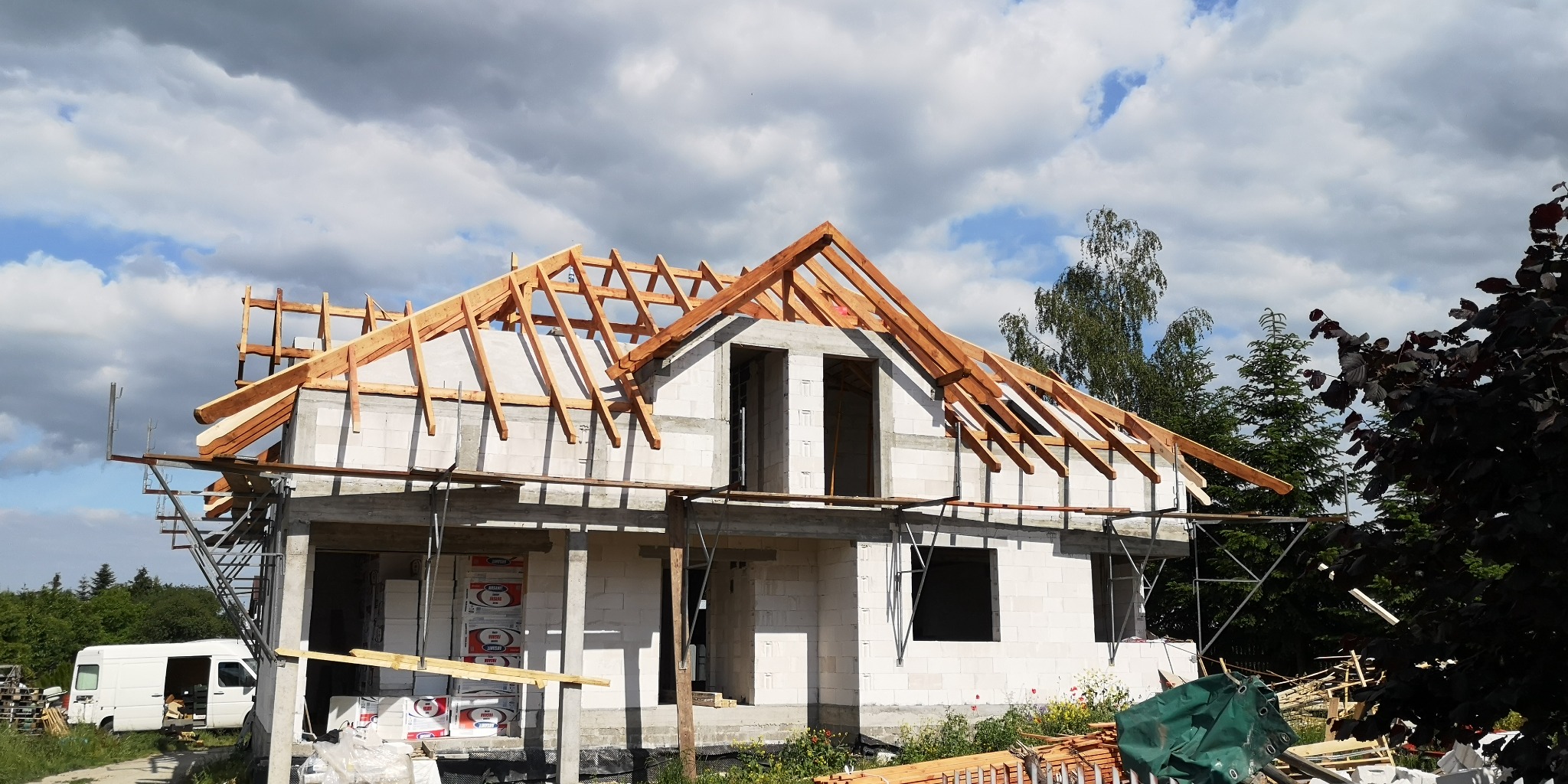 Budowa domu jednorodzinnego w stanie surowym otwartym: białe bloczki, drewniana konstrukcja dachu bez pokrycia, widoczne rusztowania i materiały budowlane.