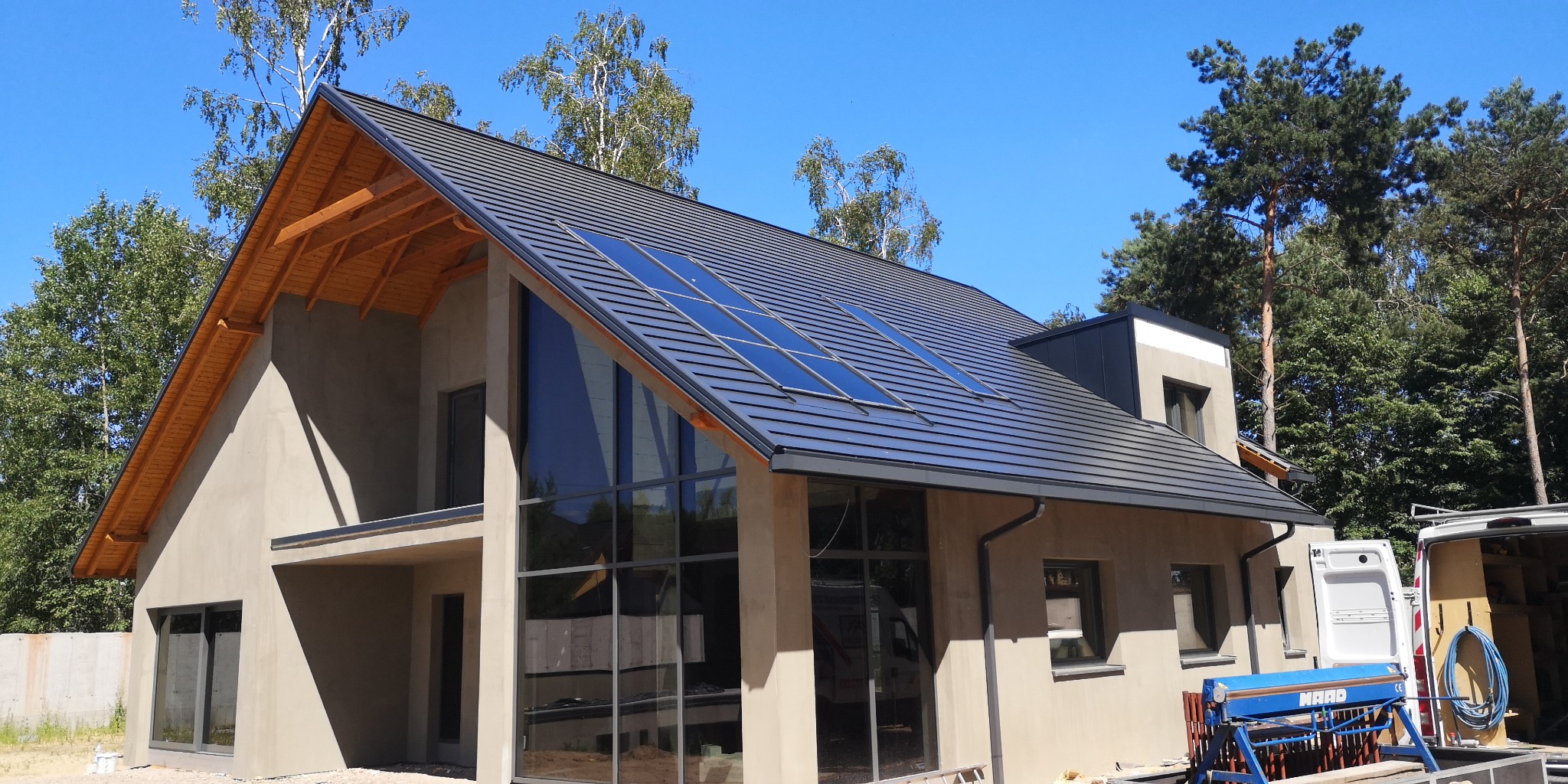 Nowoczesny dom jednorodzinny z ciemnoszarą dachówką, wbudowanymi oknami dachowymi i widoczną więźbą dachową od strony szczytu, na tle błękitnego nieba i zieleni drzew.