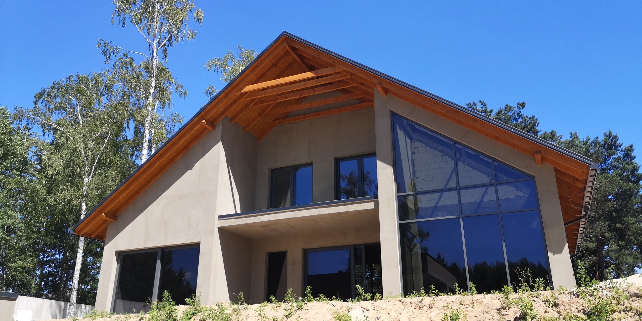 Surowy stan nowoczesnego domu z widoczną konstrukcją dachu z drewnianych belek i dużymi oknami, otoczonego drzewami i niezagospodarowanym terenem.