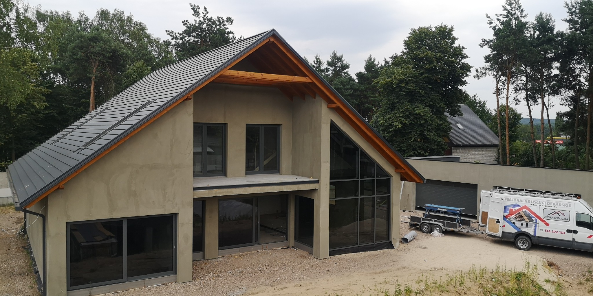 Nowoczesny dom jednorodzinny w trakcie budowy, z szarą dachówką, widoczną więźbą dachową i zaparkowanym samochodem dostawczym firmy dekarskiej.