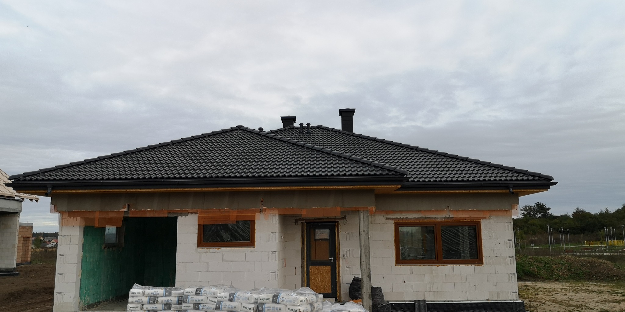 Dom w trakcie budowy z nowym, ciemnym dachem z dachówki ceramicznej, widoczne kominy i orynnowanie, worki z zaprawą murarską przed budynkiem.