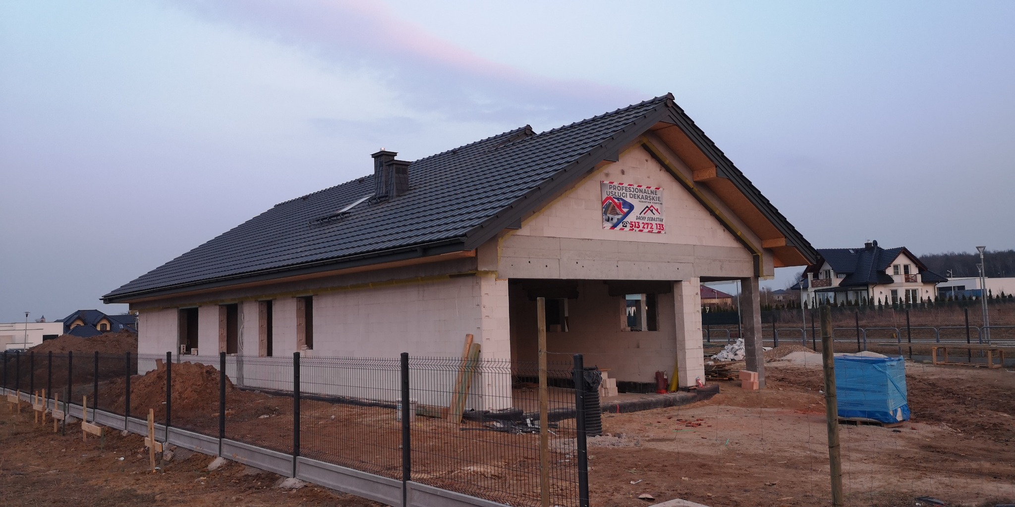 Dom w trakcie budowy z ciemnoszarą dachówką, widoczna więźba dachowa, obróbki blacharskie i komin. Na ścianie budynku baner z informacjami o usłudach dekarskich.