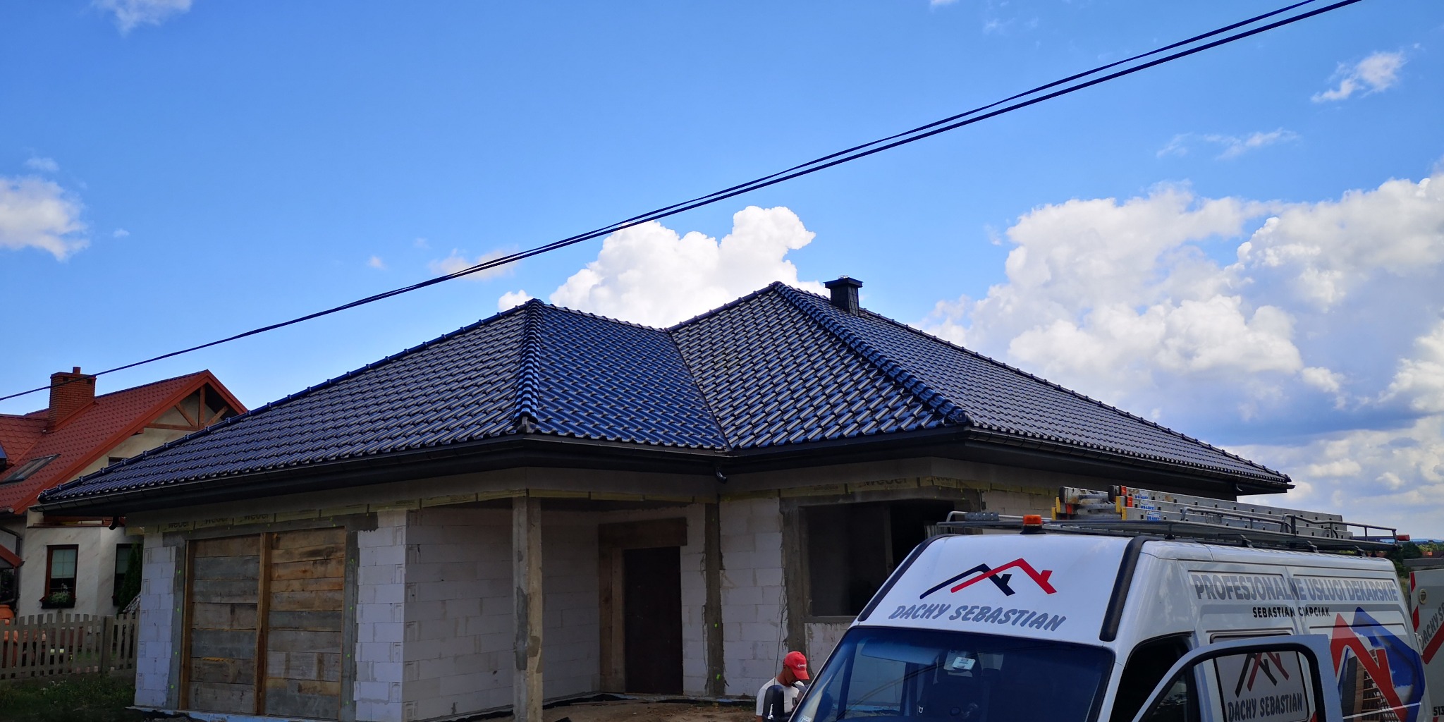 Nowoczesny dom w trakcie budowy z ciemną, ceramiczną dachówką, widoczna firma dekarska z logo na samochodzie dostawczym i rusztowaniem na dachu pojazdu.