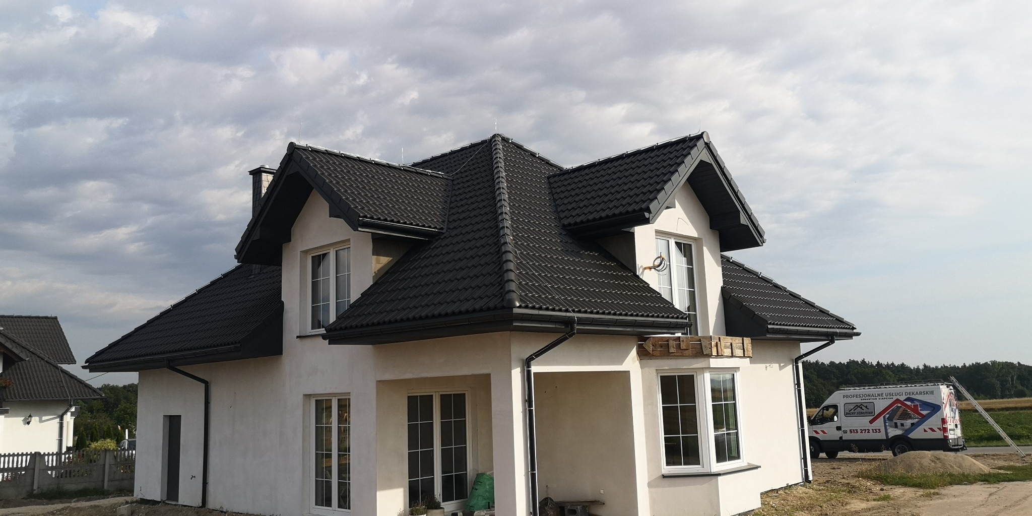 Nowoczesny dom jednorodzinny z ciemną dachówką ceramiczną, widoczne rynny i obróbki blacharskie, na podjeździe zaparkowany bus firmy dekarskiej.