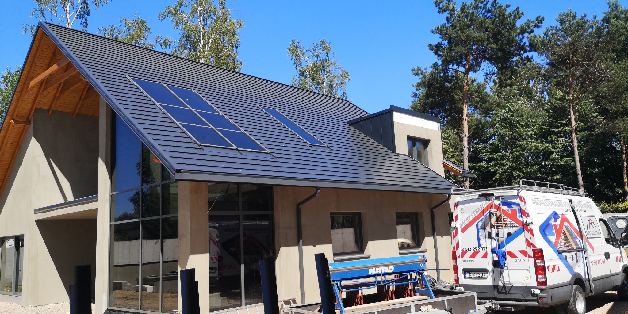 Nowoczesny dom jednorodzinny z ciemnoszarą dachówką ceramiczną i oknami dachowymi, widoczny fragment samochodu firmy dekarskiej i narzędzia blacharskie.