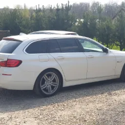 Białe BMW 5 Touring z przyciemnionymi szybami bocznymi i tylną, zaparkowane na żwirowym podłożu, z hakiem holowniczym. W tle widoczne drzewa i fragment budynku.