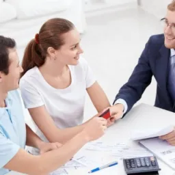 Doradca w garniturze prezentuje kartę kredytową parze przy biurku z dokumentami i kalkulatorem.