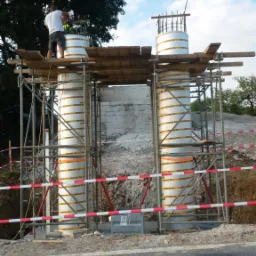 Budowa filarów mostu: dwóch robotników na rusztowaniu, betonowe kolumny w szalunkach z widocznymi zbrojeniami, zabezpieczenia placu budowy, w tle drzewa i niebo.