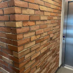 Dekoracyjna ściana z cegły klinkierowej w odcieniach czerwieni i beżu, z widocznym narożnikiem i fragmentem windy w tle.