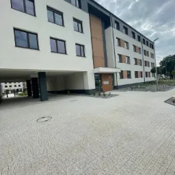 Nowoczesny, czterokondygnacyjny budynek mieszkalny z białymi ścianami, ciemnymi oknami i szarym dachem, z widocznym podjazdem wyłożonym kostką brukową i fragmentem zieleni.