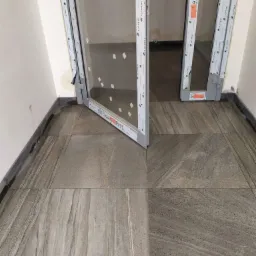 Ułożone płytki podłogowe w odcieniach szarości i beżu, z widocznym fragmentem ramy okiennej i niskim progiem, ukazujące wykończenie wnętrza.