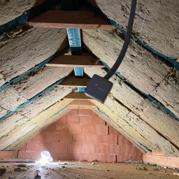 GH SYSTEMS sp. z o.o. - Ocieplenie poddasza wełną mineralną pomiędzy krokwiami, widoczne elementy konstrukcji drewnianej i fragment ściany z czerwonej cegły dziurawki, kabel zwisający z sufitu.