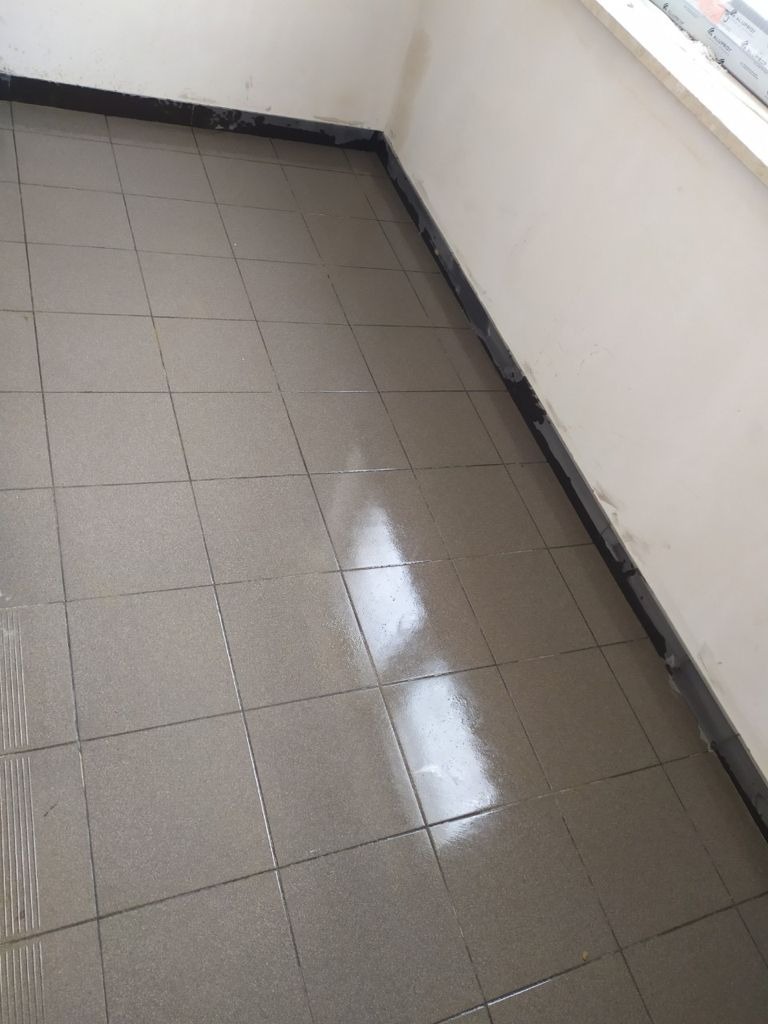 Ułożone, błyszczące, szare płytki podłogowe w małym pomieszczeniu, z ciemną listwą przypodłogową, widoczne odbicie światła na powierzchni płytek.