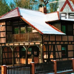 Dwukondygnacyjny dom z muru pruskiego z drewnianym balkonem i śniegiem na dachu, widoczny spod niskiego kąta, otoczony drewnianym płotem.