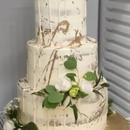 Trzypiętrowy tort weselny w stylu rustykalnym, z kremowym lukrem, złotymi plamkami i dekoracją z białych róż i zielonych liści eukaliptusa, umieszczony na złotej podkładce.
