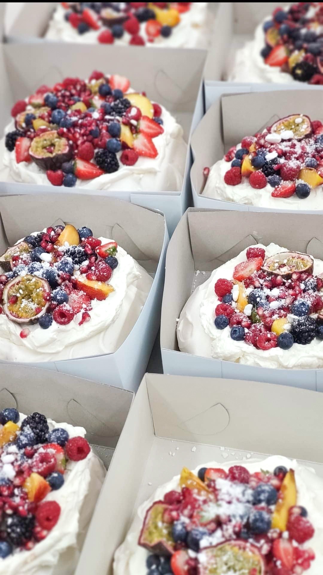 Rząd pudełek z bezowymi tortami Pavlova, każdy udekorowany bitą śmietaną i świeżymi owocami: marakują, malinami, borówkami, truskawkami, jeżynami i granatem, posypane cukrem pudrem.