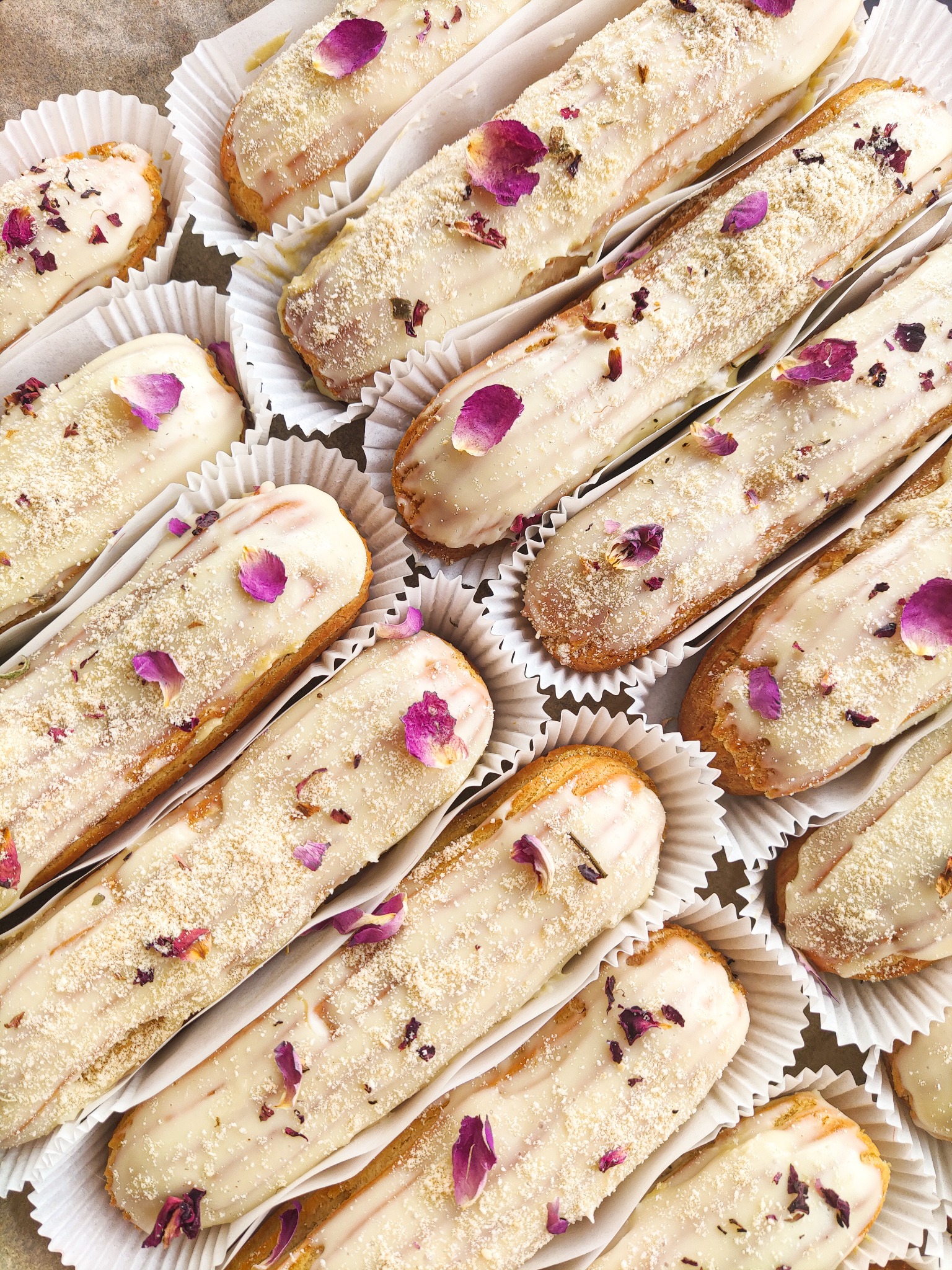 Eklery z białym lukrem, posypką i płatkami róż, ułożone w papilotkach, widok z góry.