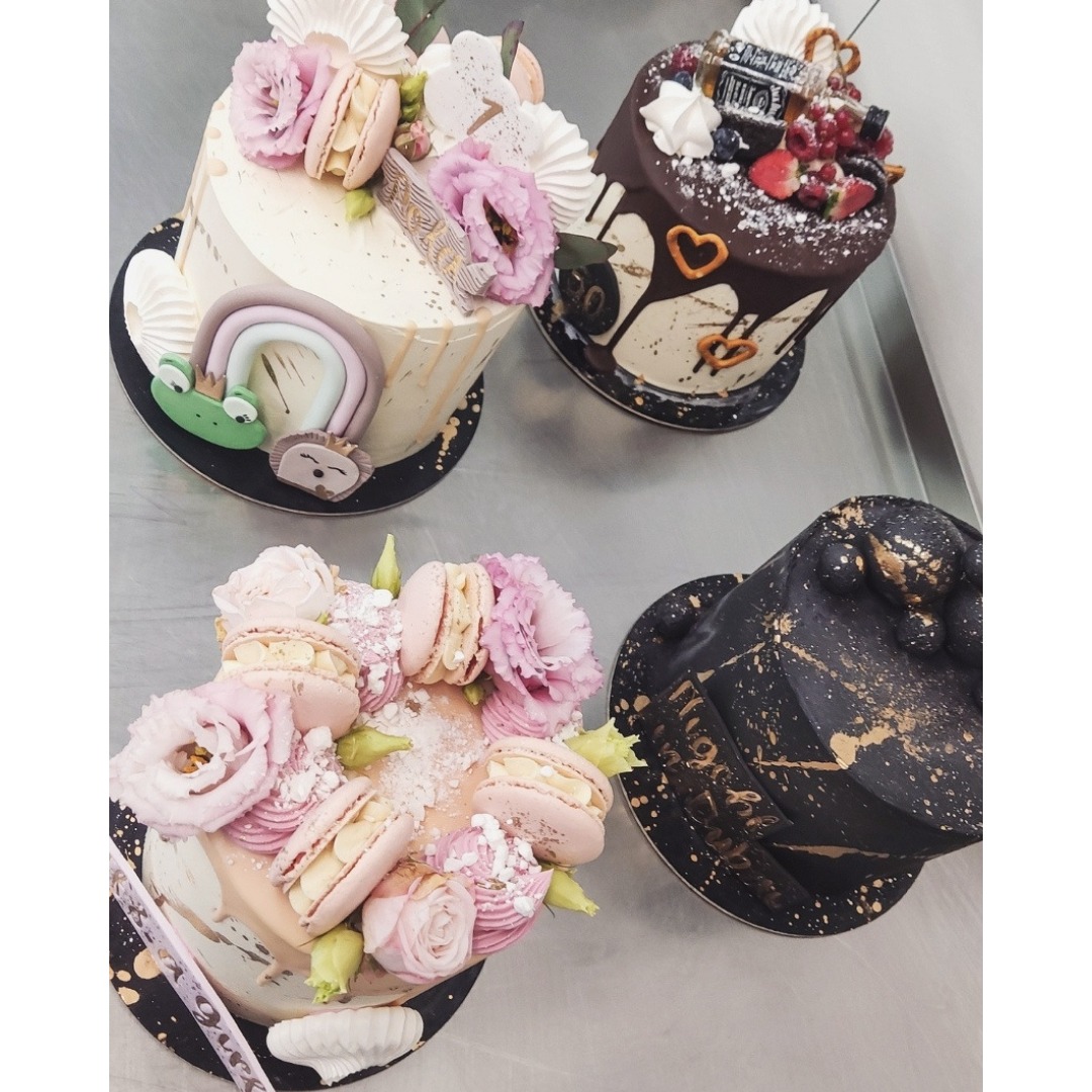 Cztery torty artystyczne na metalowym blacie: jeden z dekoracją tęczy i zwierzątek, drugi z polewą czekoladową i owocami, trzeci z różowymi kwiatami i makaronikami, czwarty czarny z napisem...