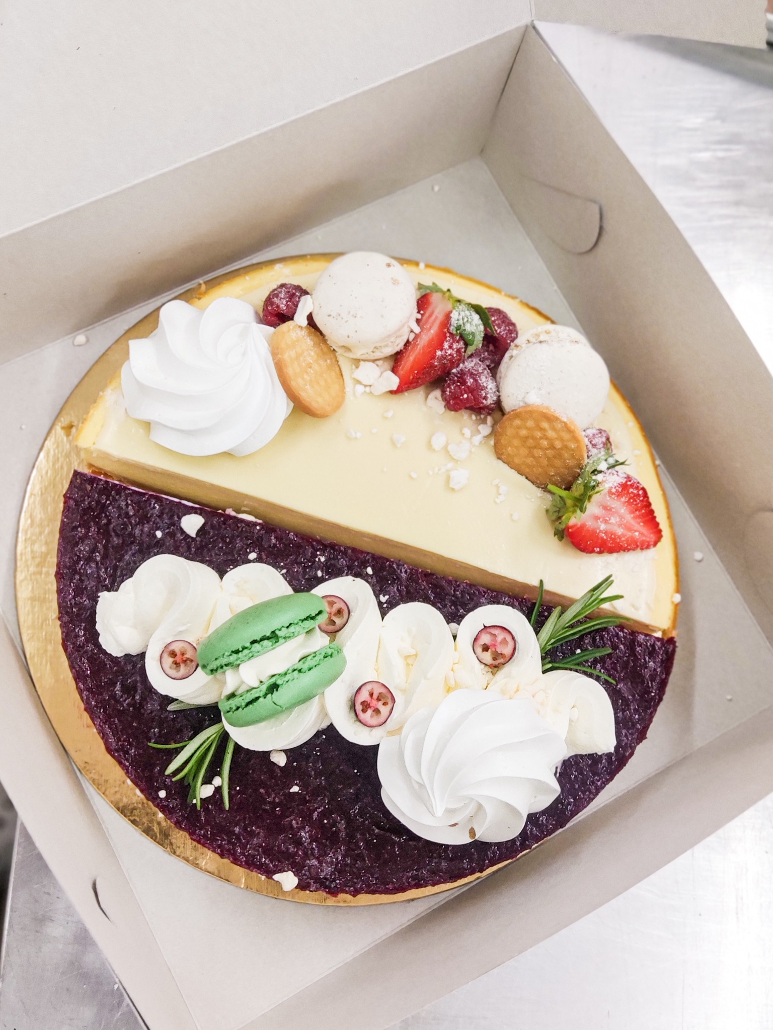 Dwukolorowy okrągły tort w kartonowym pudełku, podzielony na połowy: kremowy z bitą śmietaną, malinami, truskawkami i makaronikami oraz fioletowy z musem jagodowym, bitą śmietaną, rozmarynem...