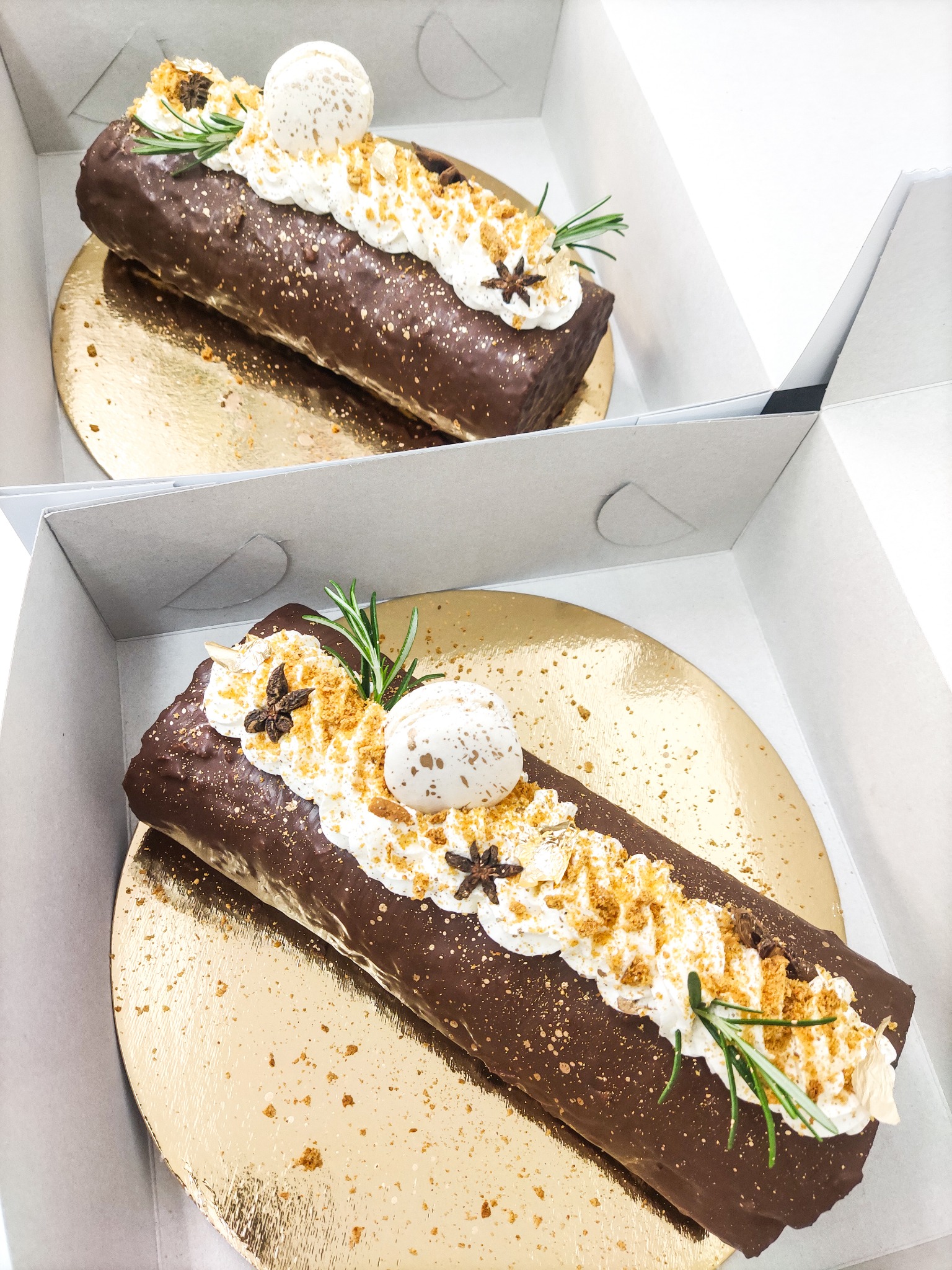 Dwa czekoladowe ciasta w kształcie pni drzewa, udekorowane bitą śmietaną, posypką, anyżem gwiaździstym i gałązkami rozmarynu, umieszczone na złotych podkładkach w białych kartonowych pudełkach.