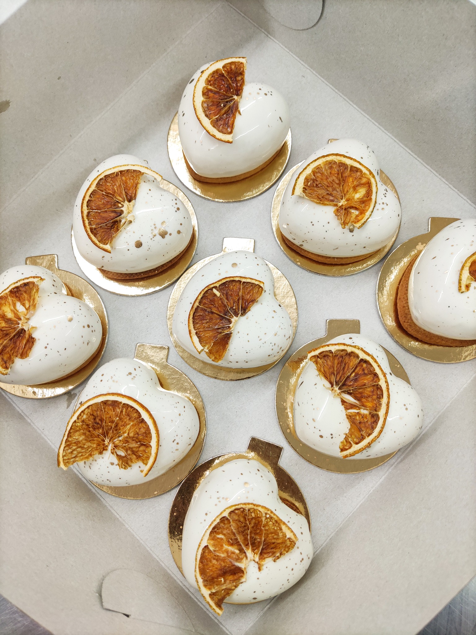 Osiem deserów w kształcie serca pokrytych białą glazurą z dekoracją z suszonej pomarańczy i złotymi kropkami, umieszczonych w kartonowym pudełku.