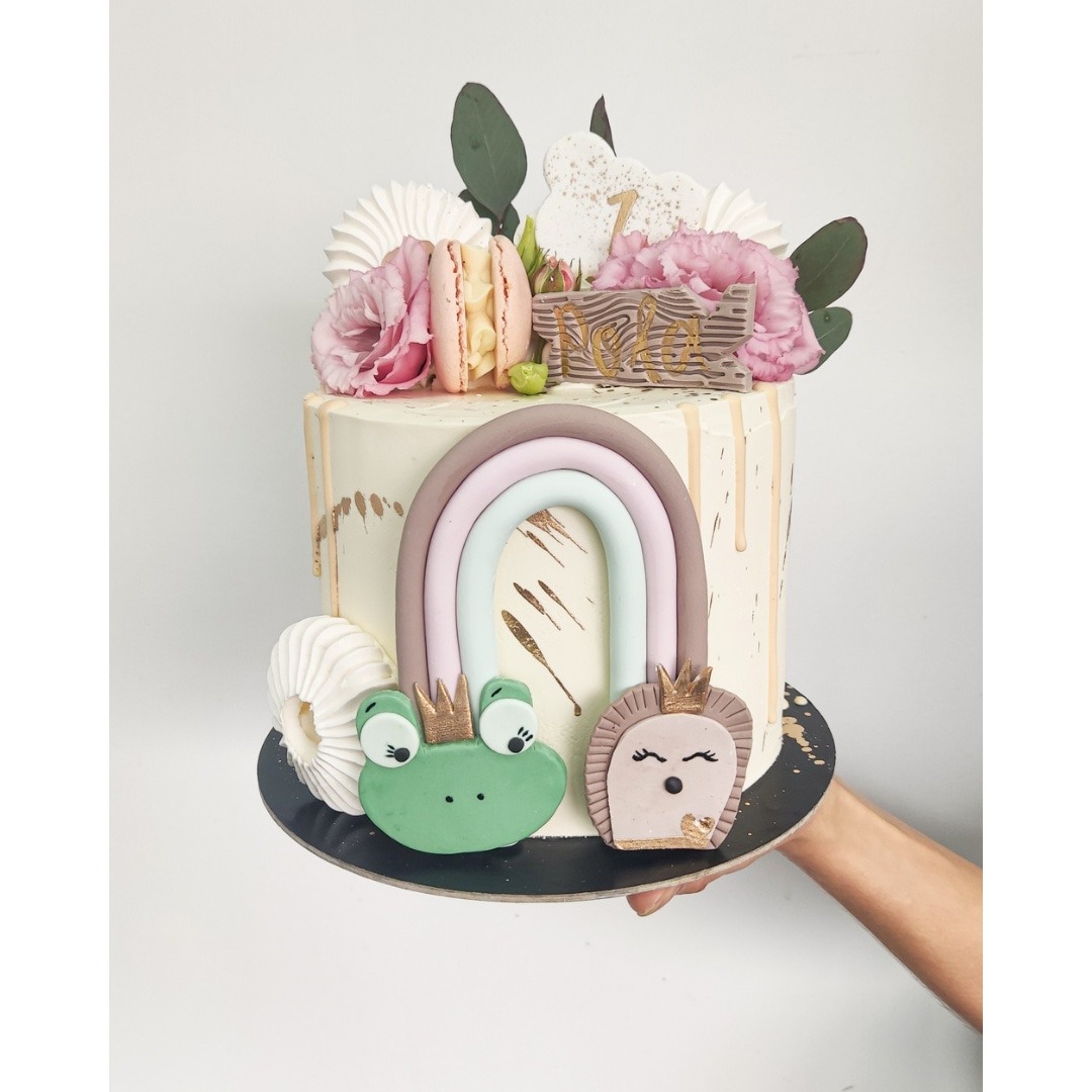 Tort z białym kremem, dekorowany tęczą, figurkami żaby i jeża w koronach, makaronikiem, bezami, różowymi kwiatami i drewnianą tabliczką z napisem, trzymany w dłoni na czarnym podeście.