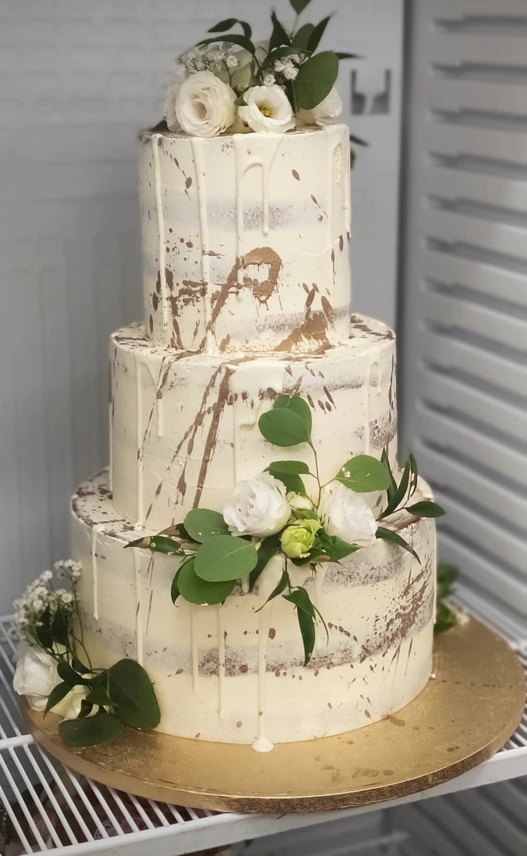 Trzypiętrowy tort weselny w stylu rustykalnym, z kremowym lukrem, złotymi plamkami i dekoracją z białych róż i zielonych liści eukaliptusa, umieszczony na złotej podkładce.