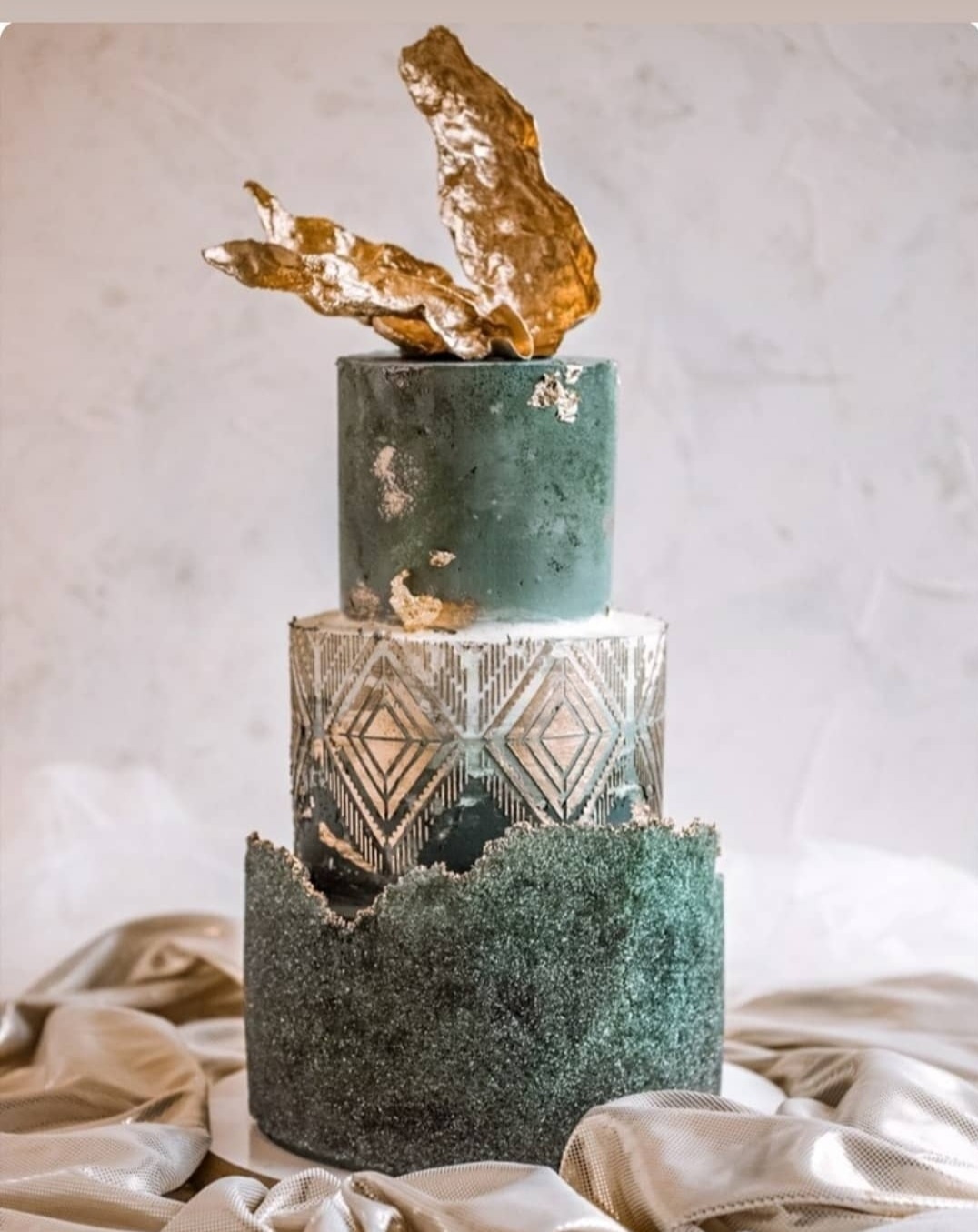 Trzypiętrowy tort w odcieniach zieleni i złota, z geometrycznymi wzorami i abstrakcyjną, złotą dekoracją na górze, ustawiony na tle drapowanej, złotej tkaniny.