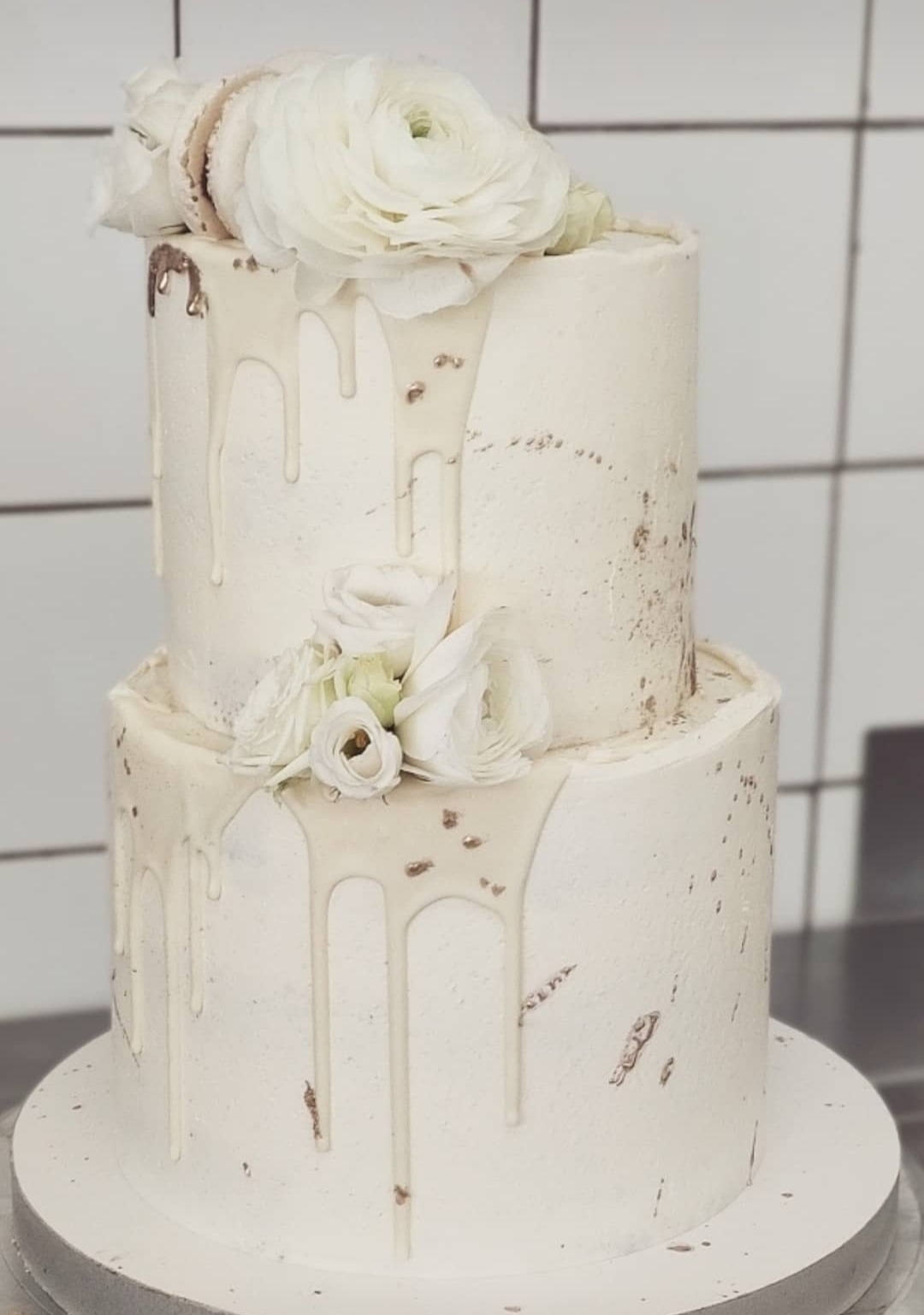 Dwupoziomowy tort weselny w odcieniach bieli, zdobiony płynącą polewą, złotymi plamkami i świeżymi, białymi kwiatami oraz makaronikami na szczycie, na tle białej ściany z płytek.