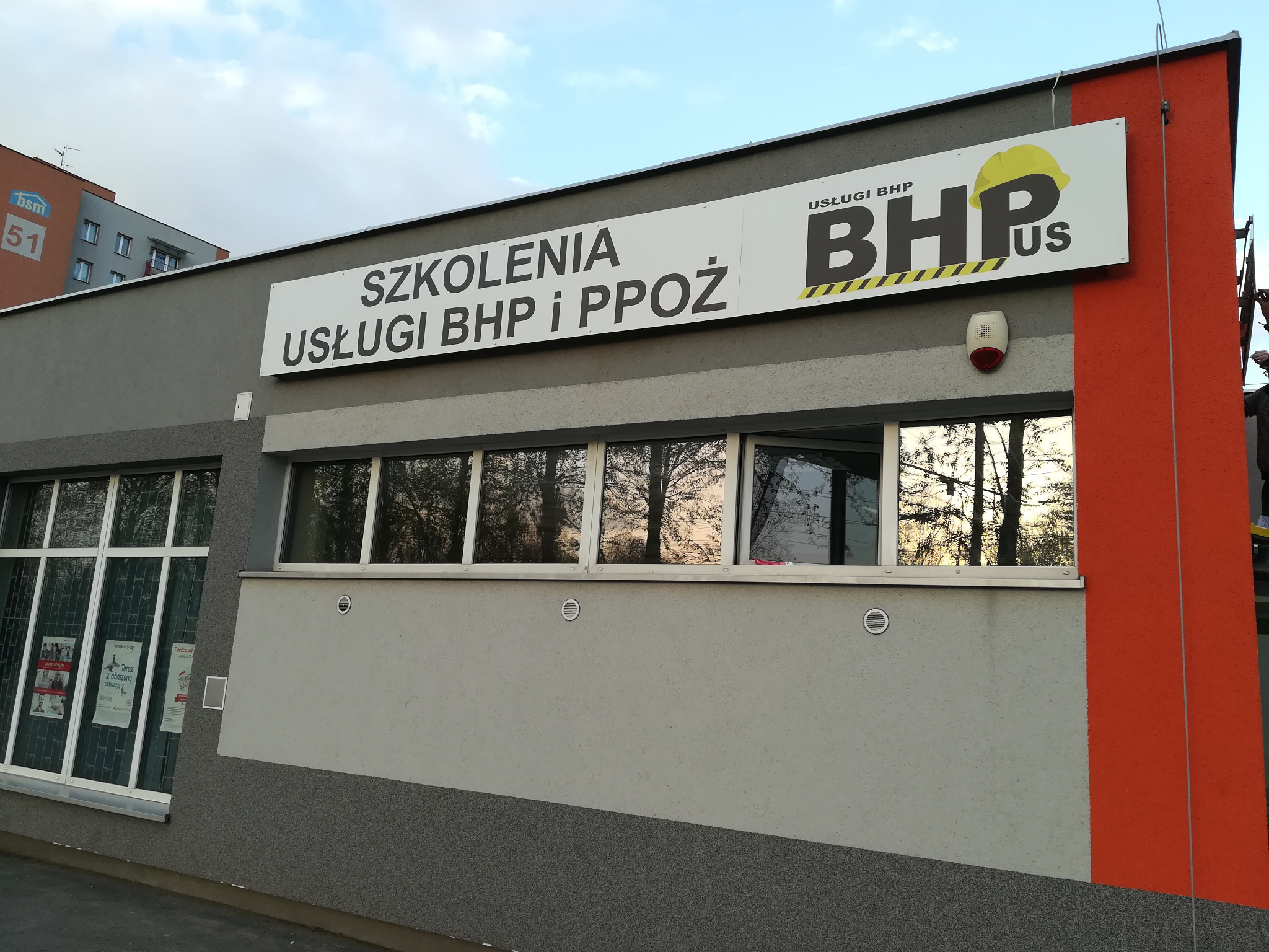 Fasada budynku z szyldami informującymi o szkoleniach i usługach z zakresu bezpieczeństwa i higieny pracy oraz ochrony przeciwpożarowej, logo firmy z żółtym kaskiem.