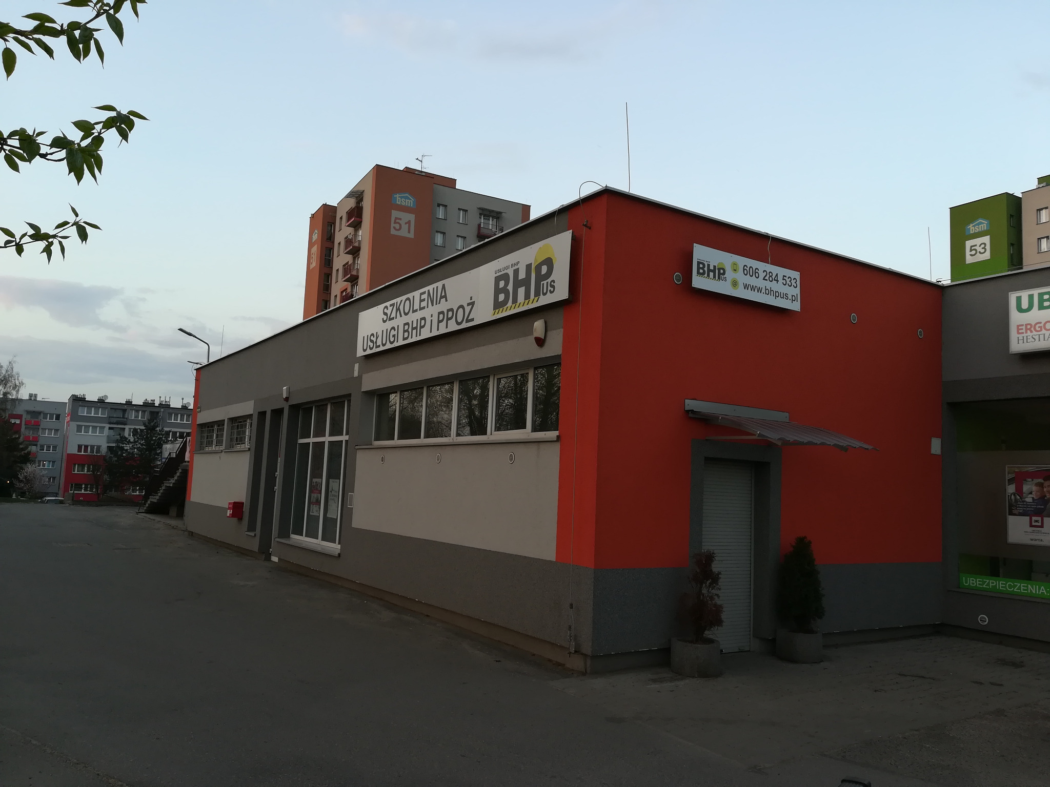 Budynek firmy oferującej szkolenia i usługi z zakresu bezpieczeństwa i higieny pracy oraz ochrony przeciwpożarowej, z widocznym logo BHP Plus i numerem telefonu na elewacji, na tle osiedla...