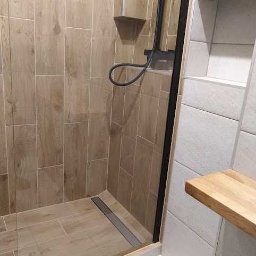 Mateusz Kędzior - Kabina prysznicowa z brązowymi płytkami imitującymi drewno, czarną armaturą i odpływem liniowym, z półkami z jasnego drewna na białej ścianie.