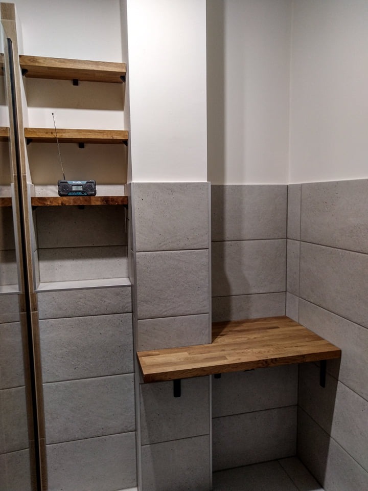 Wykończona łazienka z szarymi płytkami, drewnianymi półkami i blatem, na jednej z półek stoi radio z anteną.