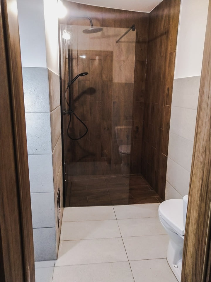 Nowoczesna kabina prysznicowa typu walk-in z drewnopodobnymi płytkami, widok z zewnątrz na białą toaletę i jasne płytki podłogowe, częściowo widoczne drewniane obramowanie drzwi.