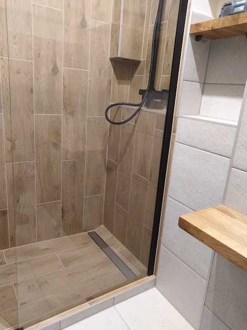 Kabina prysznicowa z brązowymi płytkami imitującymi drewno, czarną armaturą i odpływem liniowym, z półkami z jasnego drewna na białej ścianie.