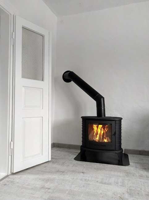 Czarny piec wolnostojący z płomieniami w minimalistycznym, białym wnętrzu z drewnopodobną podłogą i częściowo otwartymi białymi drzwiami.