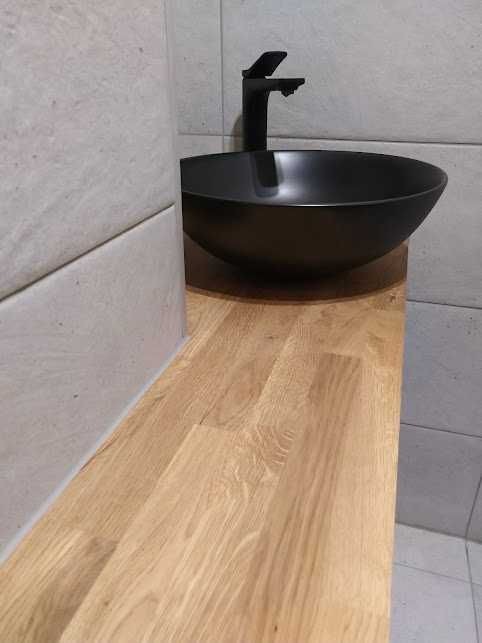 Czarna, okrągła umywalka nablatowa z czarną baterią na blacie z jasnego drewna, na tle płytek w odcieniach szarości.